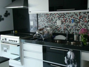 Uma cozinha moderna planejada com esquema de cores preto e branco apresenta backsplash de azulejos com mistura de azulejos vermelhos, brancos e pretos. Há um fogão, uma pia dupla com pratos em um escorredor e um vaso de flores rosa no balcão. Um pano de prato está pendurado na alça de uma gaveta.