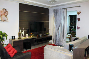 Uma moderna sala de estar com TV de tela plana montada na parede e painéis de madeira escura também funciona como um home theater aconchegante. Um sofá bege e uma poltrona cinza com almofadas vermelhas ficam de frente para a TV. Itens decorativos, incluindo um par de figuras de girafas brancas, alinham-se no console de madeira escura. Cortinas prateadas cobrem uma grande janela.