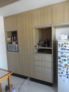 A cozinha planejada possui armários de madeira clara, geladeira branca coberta com ímãs coloridos, cafeteira preta e outros pequenos eletrodomésticos. Os armários possuem um mix de gavetas e prateleiras abertas, com micro-ondas em uma das seções abertas.