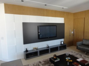 A imagem mostra uma sala de estar moderna com uma TV de tela plana montada em um painel preto contra uma parede com detalhes em branco e madeira, criando uma sensação refinada de home theater. Abaixo da TV, há um elegante console de mídia cinza contendo itens decorativos. Uma poltrona estampada cinza é visível à direita.