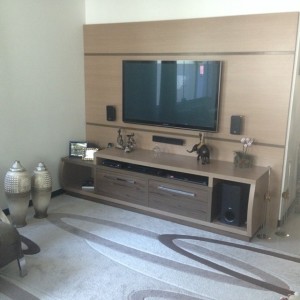 Uma moderna sala de estar dispõe de uma TV de tela plana montada na parede acima de um centro de entretenimento de madeira, com alto-falantes de home theater em ambos os lados. O armário contém vários itens, incluindo uma pequena prateleira com decoração. Dois vasos decorativos são colocados no chão à esquerda.