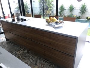 A moderna cozinha planejada possui bancada branca e armários de madeira, com porta frutas com frutas variadas. O piso apresenta azulejos com padrões complexos, enquanto grandes janelas de vidro oferecem vista para um exuberante quintal com vegetação e uma piscina.