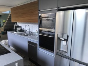 Uma cozinha moderna planejada com eletrodomésticos de aço inoxidável, incluindo forno duplo, geladeira francesa com dispensador de água e lava-louças. Há uma pia elegante com torneira, armários de madeira e cinza e um backsplash de azulejos geométricos. Uma escada é visível ao fundo.