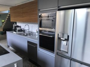 Cozinha moderna com eletrodomésticos em inox: frigorífico de duas portas com dispensador de água, forno embutido e bancada com lavatório e torneira. A cozinha planejada possui um armário de parede em madeira acima. Ao fundo, é visível uma escada. O chão tem azulejos estampados.