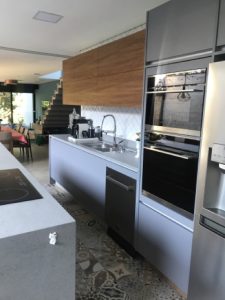Cozinha moderna com elegantes armários cinza, forno embutido e lava-louças. Um backsplash de madeira contrasta com as bancadas cinza claro. Há uma pia embaixo da janela e um cooktop preto está instalado na ilha em primeiro plano. As escadas são visíveis ao fundo desta cozinha feita.