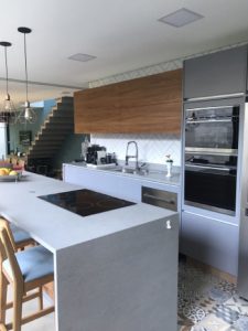 Cozinha moderna planejada com ilha cinza com cooktop de indução e banquetas de madeira. A cozinha tem armários elegantes em cinza e madeira, forno embutido e uma pia grande. A decoração inclui vasos de plantas e luminárias pendentes. Uma escada é visível ao fundo.
