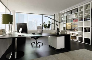 Um escritório moderno com uma elegante mesa preta, cadeira de escritório e computador desktop. Dois vasos de plantas ficam no parapeito da janela, e as grandes janelas fornecem muita luz natural. Móveis planejados incluem uma estante branca repleta de livros e itens decorativos que revestem a parede posterior.
