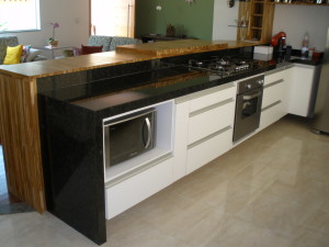 Cozinha moderna com armários brancos e bancada em granito preto. Possui fogão a gás com forno de aço inoxidável, micro-ondas embutido e balcão de bar de madeira. O fundo inclui uma área de estar com sofás, porta de vidro e decoração.