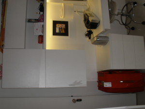 Um canto de uma cozinha moderna com armários brancos, bancadas e um design elegante. Há uma luminária branca, uma cafeteira e uma foto emoldurada no balcão. Uma mala vermelha está no chão. O espaço é iluminado com uma iluminação suave e quente.