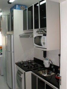 Uma cozinha moderna com bancada em granito preto, armários brancos e eletrodomésticos em aço inoxidável. Há um fogão com exaustor, um micro-ondas acima e uma geladeira branca à esquerda. Além disso, há um liquidificador e uma cafeteira na bancada.
