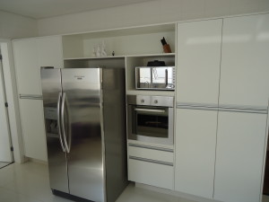 Cozinha moderna com armários brancos, geladeira side by side de aço inox, forno embutido e micro-ondas. Os armários apresentam puxadores minimalistas, com prateleiras abertas acima exibindo itens decorativos. O chão e as paredes são de azulejos.