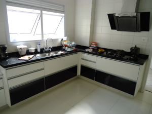 Cozinha moderna com um design elegante em preto e branco. A bancada é preta com vários itens de cozinha, incluindo liquidificador, utensílios, panelas e escorredor de pratos. O layout em forma de L apresenta fogão a gás e exaustor sob uma janela com persianas brancas.
