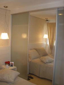 Um quarto aconchegante dispõe de cama com roupa de cama e travesseiros bege. Uma porta deslizante espelhada do armário reflete o ambiente, incluindo uma mesa de cabeceira com relógio, um pequeno porta-retratos e um vaso de flores vermelhas. As luminárias montadas na parede fornecem iluminação aconchegante e uma cortina fica pendurada na janela.