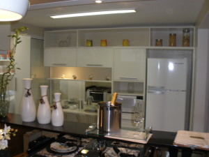 Cozinha moderna com armários brancos e eletrodomésticos encastrados, incluindo forno e frigorífico. Uma bancada apresenta vasos brancos decorativos, uma garrafa de champanhe em um balde de gelo com taças e talheres bem organizados. As prateleiras exibem potes e itens de cozinha.