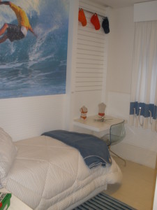 Um quarto com tema de surf apresenta um mural de um surfista pegando uma onda em uma parede. A cama é coberta com um edredom branco e um cobertor azul. Uma escrivaninha com uma cadeira transparente e vários gadgets fica ao lado da janela com cortinas brancas. Chapéus estão pendurados na parede acima.