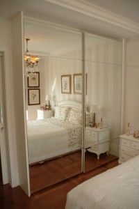 Um quarto com um grande guarda-roupa espelhado refletindo o ambiente. No interior, as paredes apresentam faixas verticais de cor clara, sendo visível uma cama com colcha floral. Há gravuras botânicas emolduradas na parede e duas mesinhas de cabeceira brancas com itens decorativos e luminárias.