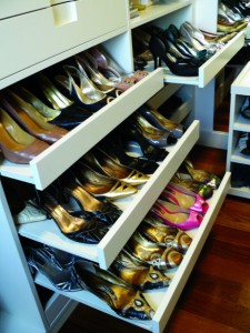 Um armário bem organizado com três sapateiras extraíveis cheias de vários pares de sapatos femininos de salto alto em diferentes cores e estilos. As estantes e prateleiras circundantes são brancas e o chão é de madeira.