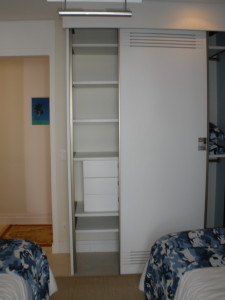 Armário com porta de correr parcialmente aberta revelando prateleiras e gavetas no interior. Duas camas de solteiro com lençóis estampados em azul e branco estão em cada lado do armário. O quarto tem tons neutros e um corredor de entrada com uma pequena pintura de uma palmeira.