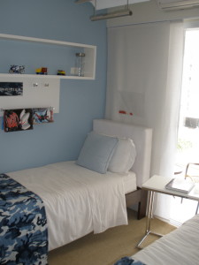 Um quarto com paredes azuis claras dispõe de duas camas de solteiro com roupa de cama branca e cobertores camuflados azuis. Cada cama tem um travesseiro branco e uma almofada azul clara. Acima das cabeceiras há prateleiras com itens decorativos. Ao lado das camas, há uma mesinha branca perto de uma janela com cortinas transparentes.
