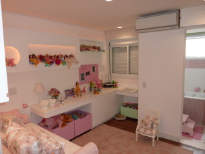 Um quarto de criança bem organizado com tema rosa. Bonecos e bichos de pelúcia ficam expostos nas prateleiras. Uma mesa com uma luminária e vários brinquedos fica embaixo de uma janela com persianas. Uma pequena cadeira de balanço e uma caixa de brinquedos estão presentes. Um aparelho de ar condicionado é montado acima da janela.