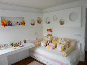 Um quarto infantil bem iluminado apresenta um sofá-cama branco adornado com almofadas estampadas e florais rosa e amarelas. Prateleiras embutidas acima e ao redor da cama exibem vários bichinhos de pelúcia e brinquedos coloridos. Uma mesa branca com mais brinquedos é colocada ao lado da cama.
