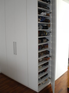 Um armário alto e branco com várias prateleiras abertas exibindo sapatos bem organizados. O gabinete tem um design moderno com alças verticais elegantes. O fundo apresenta piso de madeira e parede branca lisa.