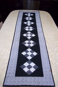 Um longo corredor de mesa retangular acolchoado com fundo preto e padrões geométricos de diamantes brancos ao longo do centro. O corredor de mesa apresenta uma borda floral branca e é colocado sobre uma toalha de mesa bege.