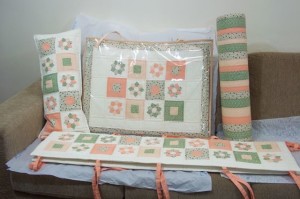 Uma coleção de itens acolchoados feitos à mão em tons pastéis suaves, apresentando uma mistura de padrões de pêssego, verde e branco. O conjunto inclui travesseiro, colcha, passadiço e almofada enrolada, todos adornados com desenhos florais e geométricos em patchwork, dispostos sobre um sofá bege.