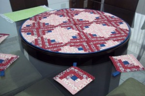 Uma mesa de jantar redonda é coberta por uma colcha circular com padrão geométrico vermelho e branco com quadrados azuis marinho. Bases para copos quadradas correspondentes são colocadas ao redor da mesa, e um pano verde é parcialmente visível no fundo.