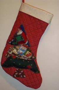 Uma meia de Natal vermelha com bolinhas brancas, adornada com uma aplicação de tecido de um Papai Noel e um desenho de árvore de Natal, está pendurada na parede. A meia tem a parte superior debruada em branco e uma alça para pendurar.