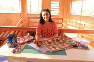Uma mulher está sentada à mesa com vários materiais para quilting, incluindo quadrados de tecido, um cortador rotativo e outros materiais de costura. Ela sorri, vestindo um suéter vermelho com bolinhas pretas. O fundo possui amplas janelas e acabamentos em madeira, sugerindo um espaço bem iluminado e aconchegante.
