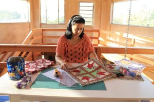 Uma mulher está sentada à mesa trabalhando em uma colcha em uma sala iluminada pelo sol e com grandes janelas. Ela tem ferramentas de quilting, tecidos e uma lata sobre a mesa. Ela está focada em seu trabalho, cercada pelo interior aconchegante de madeira da sala.