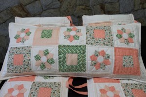 Conjunto de almofadas decorativas com desenhos em patchwork com padrões florais e geométricos em tons de rosa, verde e creme. As almofadas estão dispostas em camadas contra o fundo de uma parede de pedra.