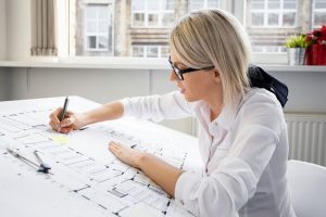 Uma mulher loira e de óculos está sentada em uma mesa, trabalhando atentamente em projetos arquitetônicos para uma marcenaria em um escritório bem iluminado. Ela está segurando uma caneta e parece estar desenhando ou fazendo anotações nos planos. As janelas ao fundo revelam um prédio de tijolos.