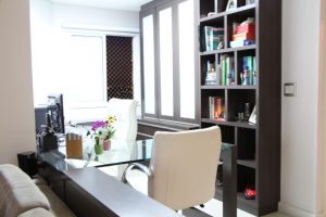 Escritório doméstico moderno com mesa de vidro, cadeira de couro branco e estantes de madeira escura repletas de livros e decoração. Um computador está sobre a mesa, junto com algumas pequenas plantas e material de escritório. A luz entra por uma janela com vista para o exterior de um edifício, mostrando a beleza dos escritórios domésticos sob medida.