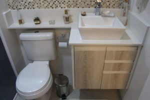 Esta imagem mostra um banheiro compacto e moderno. Possui um vaso sanitário branco adjacente a uma pia retangular branca colocada sobre uma penteadeira com armários de madeira clara. Acima da pia há um backsplash de mosaico decorativo. Vários produtos de higiene pessoal estão cuidadosamente dispostos na pia e na prateleira, ecoando a elegância dos banheiros sob medida.