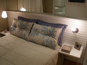 Um quarto aconchegante com uma colcha bege na cama. Duas almofadas grandes, cada uma com um desenho botânico, ficam encostadas na cabeceira da cama. Duas pequenas lâmpadas de cada lado da cama proporcionam uma iluminação suave. Uma mesa de cabeceira branca contém uma pequena caixa e uma foto emoldurada, tudo parte de dormitórios sob medida habilmente projetados.