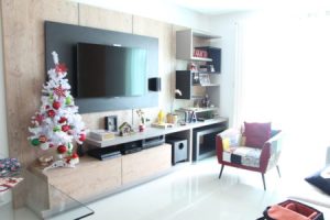 Uma moderna sala de estar com TV de tela plana montada acima de uma unidade de entretenimento de madeira, criando a experiência perfeita de home theater. Uma árvore de Natal branca com enfeites vermelhos e verdes fica próxima. A sala inclui uma cadeira estofada multicolorida, prateleiras com decorações e uma janela iluminada ao fundo.