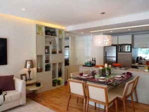Interior moderno do apartamento com layout em plano aberto. A área de estar possui sofá branco com almofadas, piso de madeira clara e mesa lateral com abajur. A área de jantar inclui uma mesa de vidro com capacidade para seis pessoas sob uma luminária suspensa. Os armários personalizados da marcenaria em São Caetano valorizam os eletrodomésticos e utensílios de inox da cozinha.