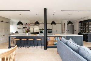 Uma moderna cozinha e área de jantar com duas grandes ilhas com bancos de bar, iluminação pendente e prateleiras embutidas. Armários neutros complementam as bancadas escuras e elegantes. Em primeiro plano, um sofá azul e uma mesa de jantar de madeira são parcialmente visíveis.