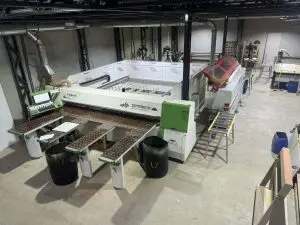 Uma oficina industrial com configuração de máquina de corte a laser Trotec Speedy. Vários materiais e equipamentos estão disponíveis, incluindo barris, mesas e outras máquinas. A área possui piso de concreto e vigas metálicas de sustentação.