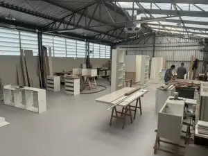 Uma grande oficina com diversas pessoas trabalhando na montagem de móveis brancos. Vários móveis como armários, gavetas e bancadas estão espalhados pelo espaço. A sala tem teto alto com treliças metálicas e ampla iluminação natural pelas janelas.