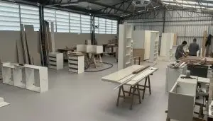 Uma oficina espaçosa com pé direito alto e ampla iluminação. Os trabalhadores estão montando diversos móveis, incluindo prateleiras e armários. O chão está bem organizado com ferramentas e materiais, e as paredes possuem grandes janelas que proporcionam luz natural.