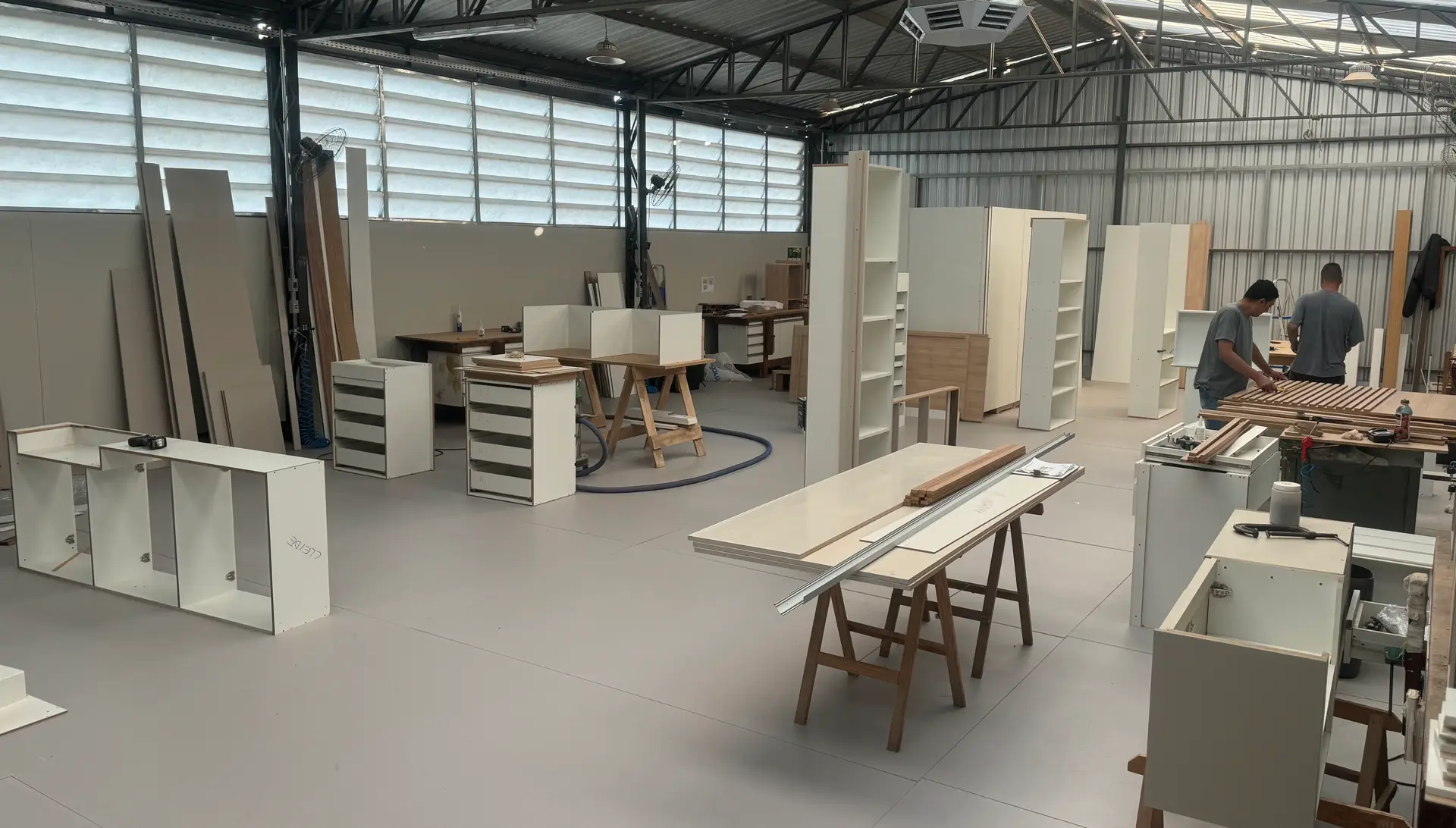 Uma oficina espaçosa com pé direito alto e ampla iluminação. Os trabalhadores estão montando diversos móveis, incluindo prateleiras e armários. O chão está bem organizado com ferramentas e materiais, e as paredes possuem grandes janelas que proporcionam luz natural.
