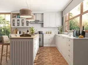 Cozinha moderna com armários brancos, grande ilha com bancada e banquetas de madeira e piso de cerâmica hexagonal. O espaço é iluminado por grandes janelas com vegetação exterior visível. Luzes pendentes estão penduradas acima da ilha e vários itens de cozinha estão bem organizados.
