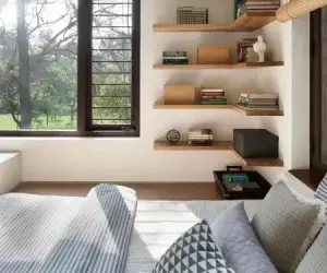 Um quarto aconchegante com cama bem feita e roupa de cama estampada em cinza e branco. Prateleiras flutuantes de madeira adornadas com livros e itens decorativos são montadas na parede. A luz solar entra através de grandes janelas, oferecendo uma vista de uma paisagem verdejante no exterior.